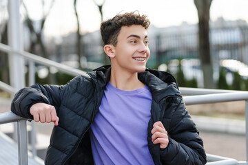 Smiling confident millennial boy, teenager with dental braces wearing black jacket looking away