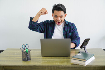 Excited screaming young asian man looking at the screen of his laptop and triumphing with raised hands