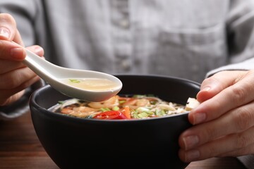 Woman eating delicious ramen with spoon at table, closeup. Noodle soup