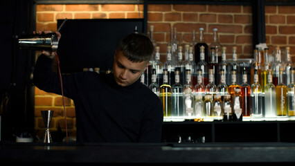 The barman preparing perfect cocktail, standing behind the bar counter. Media. Many bottles of alcohol on the background. - obrazy, fototapety, plakaty
