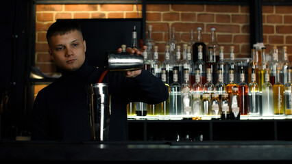 The barman preparing perfect cocktail, standing behind the bar counter. Media. Many bottles of alcohol on the background. - obrazy, fototapety, plakaty