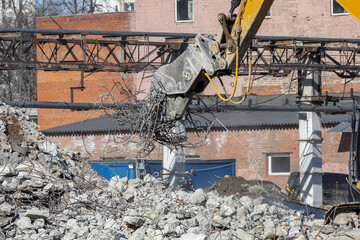 Fototapeta premium A yellow excavator with a hydraulic concrete pulverizer lifts a pile of old metal reinforcing bars over a pile of concrete rubble left over from the demolition of an old building. Close-up