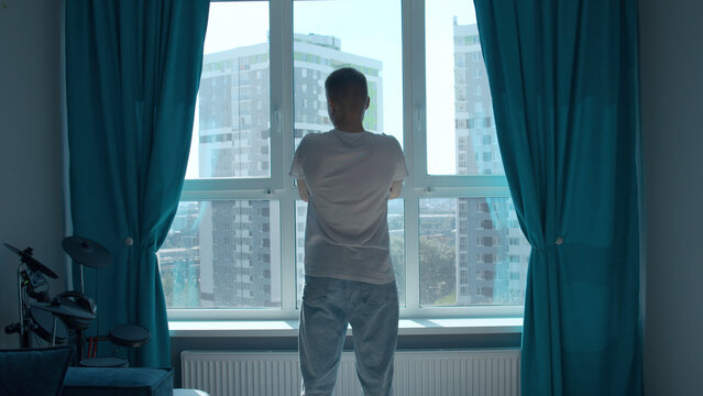 Rear view of a pensive man standing at home by the window and looking at the city. Media. Man in home clothes spending time in reflection in his bedroom.
