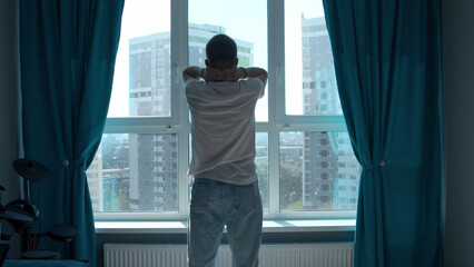 Early morning, silhouette of man exercising, stretching body by window at home. Media. Rear view of a young man standing against window.