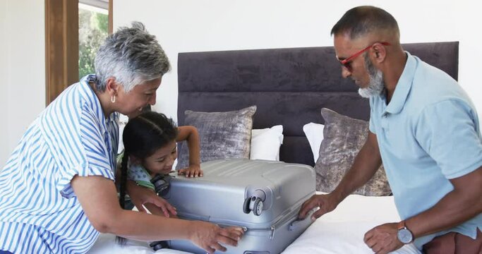 Biracial grandparents with their granddaughter unpack a suitcase on a bed
