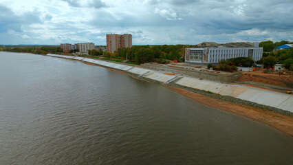 Fototapeta premium Aerial view of city embankment with construction site. Clip. Unfinished area and calm river with buildings and green trees on the background.