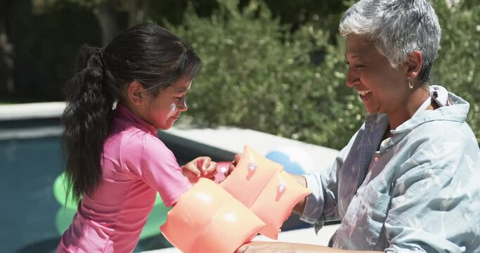 Biracial girl with a pink swimsuit plays with water wings, smiling at her grandmother