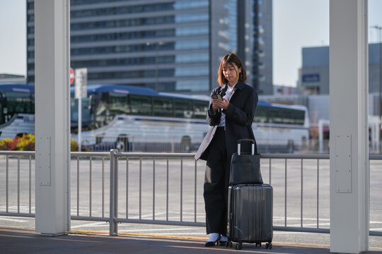 バスターミナルでスーツケースを置いてスマートフォンを見るビジネスウーマン