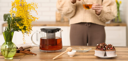 Glass teapot with hot tea, honey, sugar and cupcakes on table in kitchen