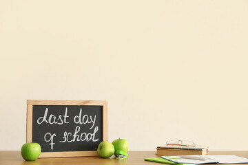 Chalkboard with text LAST DAY OF SCHOOL, apples and notebooks on table near light wall