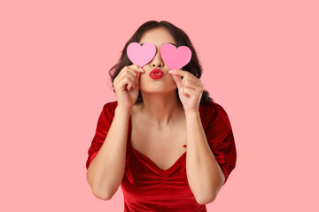 Beautiful young happy Asian woman with paper hearts blowing kiss on pink background. Valentine's Day celebration