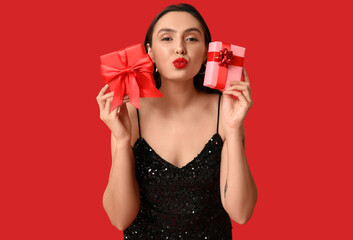 Beautiful young woman with gift boxes blowing kiss on red background. Valentine's Day celebration