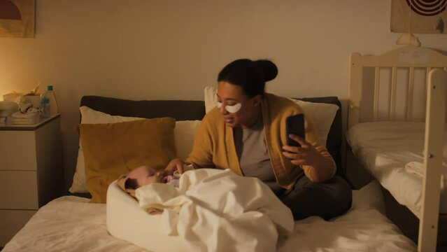 Full shot of young casually dressed African American or Hispanic woman with under eye patches sitting on bed at home, having video call on smartphone, smiling and posing together with baby