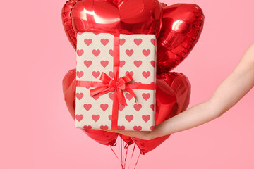 Female hand with gift box and heart-shaped balloons on pink background. Valentine's Day celebration