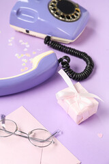 Composition with vintage telephone, gift box, eyeglasses and envelope on lilac background, closeup. Valentine's Day celebration