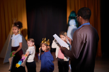 Back view of Black drama teacher directing school play with group of children on stage in theater copy space