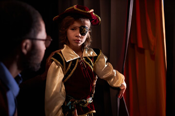 Waist up portrait of young boy wearing pirate costume standing by red curtain ready to go on stage...