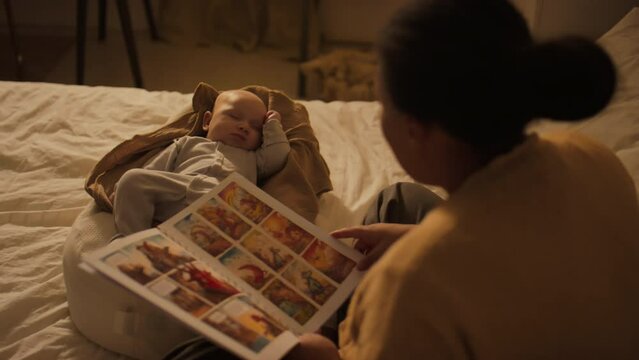 Medium over-shoulder shot of black mother sitting beside 3-month-old baby boy sleeping in nest pod, reading aloud fairy tale from colourful comic book about adventures of dinosaur family