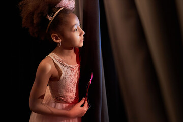 Side view portrait of African American girl wearing pink dress peeking over curtain backstage in...