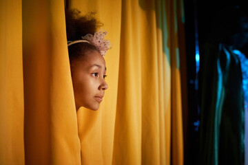 Side view portrait of African American girl peeking head on stage from yellow curtains with...