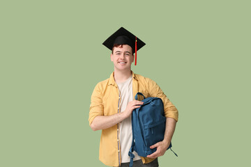 Male student in mortar board with backpack on green background