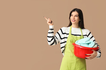 Beautiful young woman holding basket with clean laundry and pointing at something on brown background