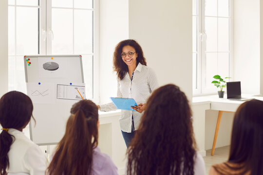 Happy young biracial woman coach in glasses with clipboard giving professional presentation in front of office board during business training class with diverse multiethnic group of people