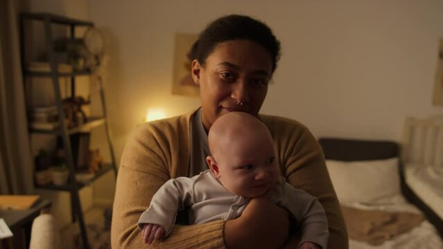 Medium portrait shot of happy young new African American mother standing in bedroom at home, swaying, holding baby son in arms and looking at camera, and boy fussing and crying