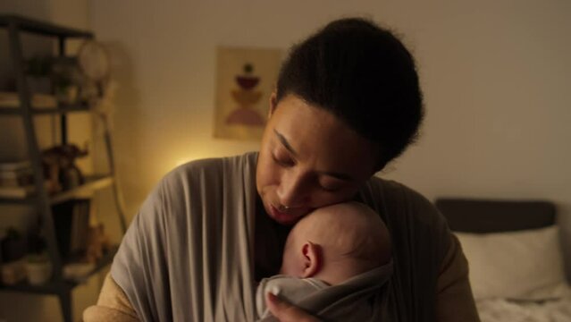 Medium close-up shot of young multiethnic woman with sleeping baby son or daughter in sling wrap walking around bedroom in daytime, swaying, cuddling and whispering lovingly
