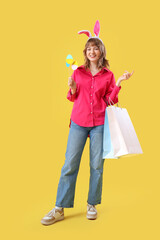 Happy young woman in bunny ears headband holding paper shopping bags and Easter decor on yellow background