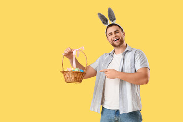 Handsome man in bunny ears pointing at Easter eggs on yellow background