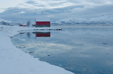 Winter landscape with red barn by the icy bay surrounded by snow covered mountains.