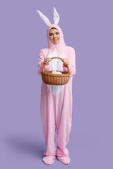 Beautiful young woman in bunny costume holding wicker basket with Easter cake and eggs on purple background