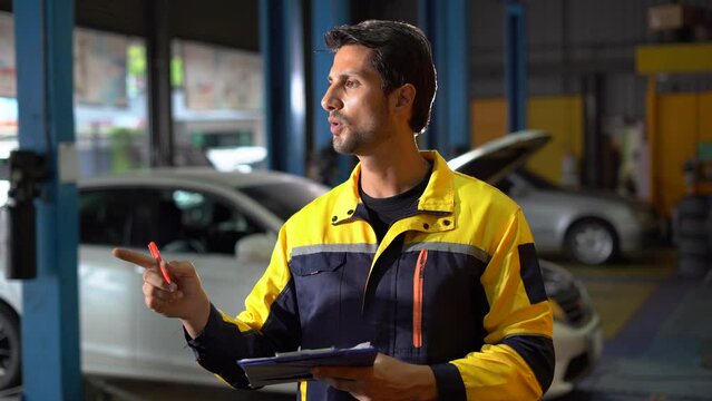 happy latin auto mechanic man checking car with clipboard gesturing show thumbs up and looking at camera in garage cars service . hispanic technician repairing vehicle at garage .