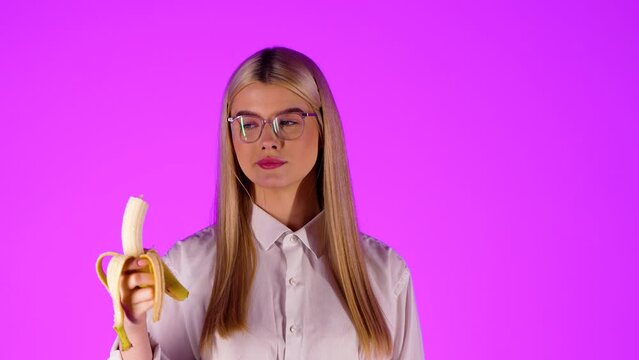 Concept oral sex, confident young woman biting a banana, pink studio background