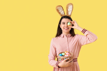 Beautiful young woman in bunny ears holding wicker basket with Easter eggs on yellow background