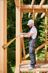 Carpenter constructing wooden skeleton building. Man measures distances with tape measure while wearing work attire and helmet. The concept of modern, environmentally-friendly construction.