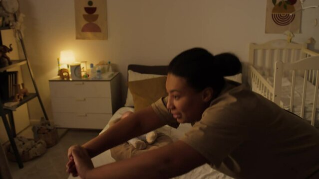 Medium shot of cheerful young body positive black mother exercising alone at home while baby son is lying in cocoon on bed - bending down, stretching, flexing, to get fit after childbirth