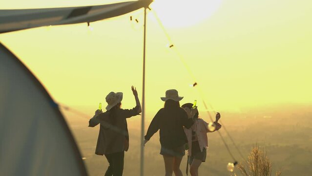 Friendship group of Asian young women having fun and enjoy partying. They are all holding bottles of beer and celebrating on the mountain outdoor camping trip in nature during sunset. slow motion