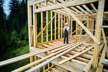 Laborer constructing wooden frame house near forest. Man treating woods, applying fire retardant...