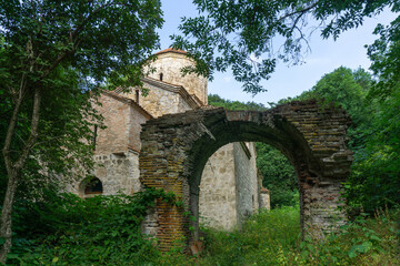Fototapeta na wymiar An old stone church with a tiled roof and a dome in the forest. Brick arch of an old wall. There are trees and bushes around. Day, bright blue sky.