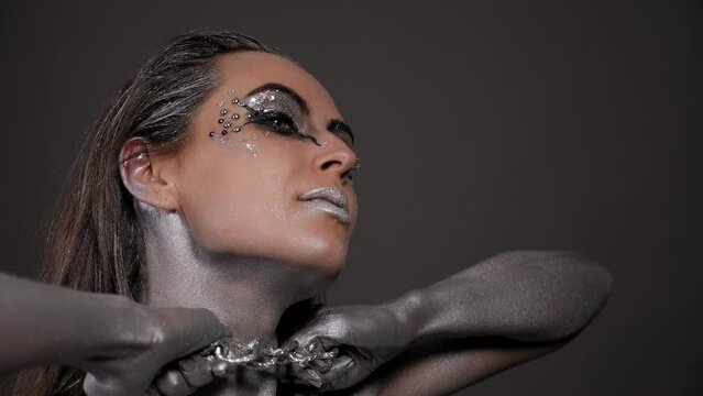 A gorgeous female model with silver body art on her body, bright makeup and silver hair poses for the camera, a chain hanging around her neck. Fashion.