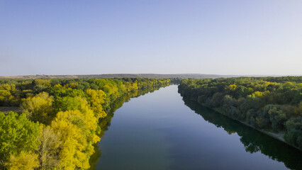 Hovering higher, the drone captures the river bordered by lush yellow-green trees, offering a...