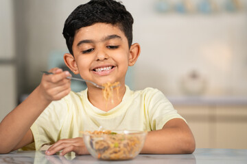 Side view shot of Indian preteen kid busy eating tasty noodles at kitchen using fork - concept of childhood fun, nutrition and enjoyment