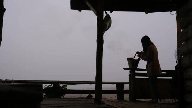 A woman lighting fire in a stove before boiling water in kettle on wooden balcony on foggy day