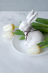 Aesthetic Easter home interior decoration, white bunny figurine on plate and tulip flowers bouquet on neutral marble table background with soft natural light