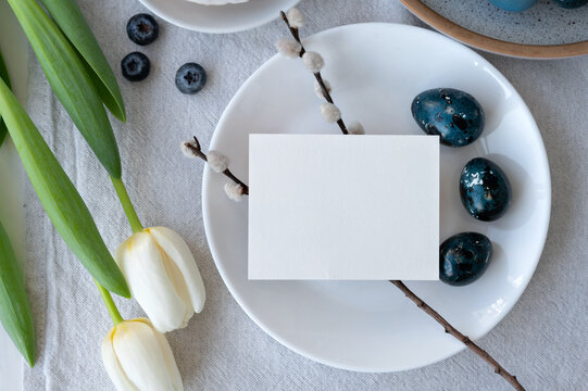 Empty paper card mockup on plate with blue dyed eggs, pussy willow and tulip flowers on neutral beige linen tablecloth, Easter holiday invitation or greeting postcard design template,