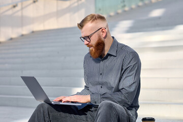 Handsome red-haired freelancer man with a beard sits on the street and works remotely using a laptop
