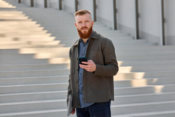 A well-groomed European man with a red beard dressed in a blue shirt and jacket against the backdrop of urban architecture