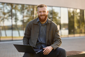 Business man with a red beard sitting on the street with an open laptop